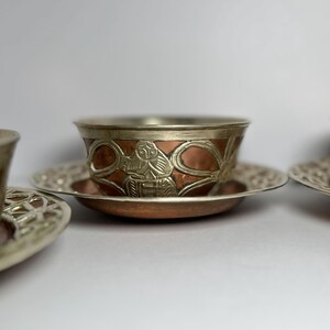 May include: Three ornate copper and silver tea cups and saucers. The cups have a copper base with silver trim and detailed relief patterns. The saucers are silver with a decorative openwork design.