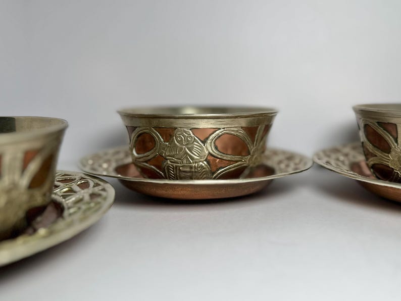 May include: Three ornate copper and silver tea cups and saucers. The cups have a copper base with silver trim and detailed relief patterns. The saucers are silver with a decorative openwork design.