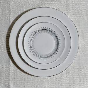 May include: Three white ceramic plates of varying sizes are stacked on top of each other. The largest plate has a silver rim, the middle plate has a decorative border, and the smallest plate is plain white. The plates are arranged on a textured white surface.