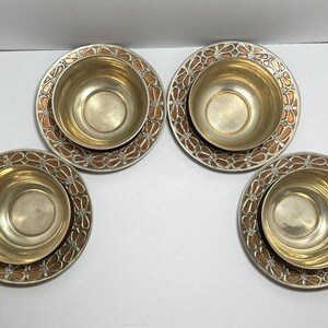 May include: Four matching metal bowls with decorative plates. The bowls are gold-colored, and the plates have a silver rim with a copper-colored floral design. The bowls sit centered on the plates.