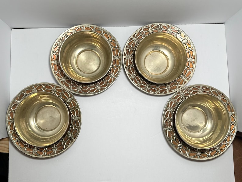 May include: Four matching metal bowls with decorative plates. The bowls are gold-colored, and the plates have a silver rim with a copper-colored floral design. The bowls sit centered on the plates.
