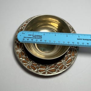 May include: A silver-colored bowl and saucer set with a floral design. The saucer has a brown and silver floral pattern around the edge. A blue ruler is placed across the bowl, measuring approximately 3 inches in diameter.
