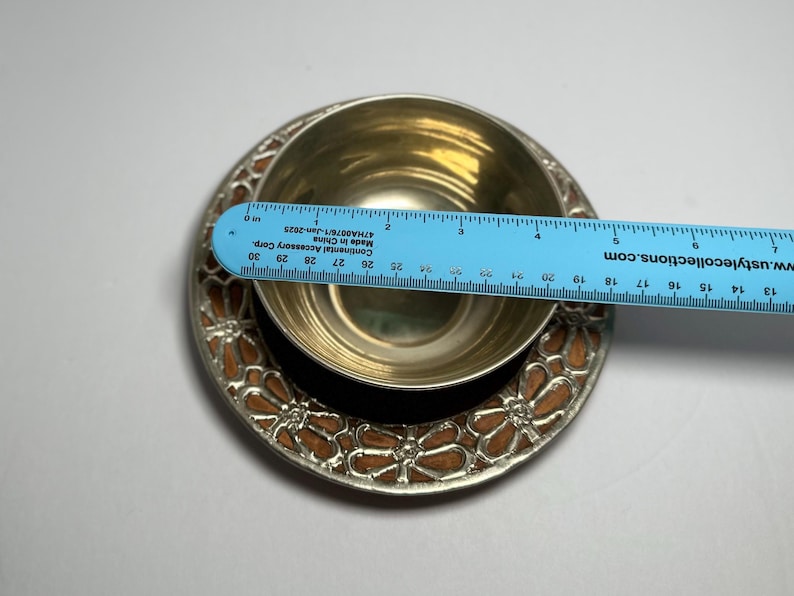 May include: A silver-colored bowl and saucer set with a floral design. The saucer has a brown and silver floral pattern around the edge. A blue ruler is placed across the bowl, measuring approximately 3 inches in diameter.
