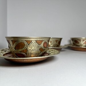 May include: Three decorative copper and silver-toned bowls and saucers. The bowls have a copper base with silver-toned rims and detailed designs. The saucers have a matching copper base and silver-toned, openwork rims. The set is displayed on a white surface.