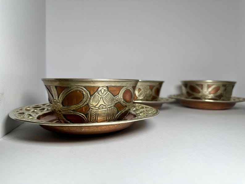 May include: Three decorative copper and silver-toned bowls and saucers. The bowls have a copper base with silver-toned rims and detailed designs. The saucers have a matching copper base and silver-toned, openwork rims. The set is displayed on a white surface.