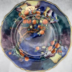 Könnte beinhalten: Ein dekorativer Teller mit gewelltem Rand und Goldrand. Der Teller zeigt ein handgemaltes Design mit Orangenfrüchten auf einem Ast vor blauem und violettem Hintergrund. Ein einzigartiges Kunstwerk.