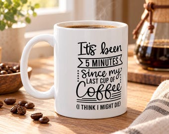 May include: A white ceramic coffee mug featuring black text that reads: It's been 5 MINUTES since my LAST CUP OF Coffee (I THINK I MIGHT DIE). The mug has a standard handle and is filled with dark brown coffee, showing a smooth, glossy finish.