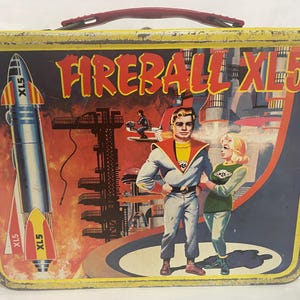 Puede incluir: Fiambrera met&aacute;lica vintage con asa roja y gr&aacute;fico colorido. El gr&aacute;fico presenta el texto "FIREBALL XL5", un cohete y dos personajes en un entorno futurista. La fiambrera tiene un borde amarillo.