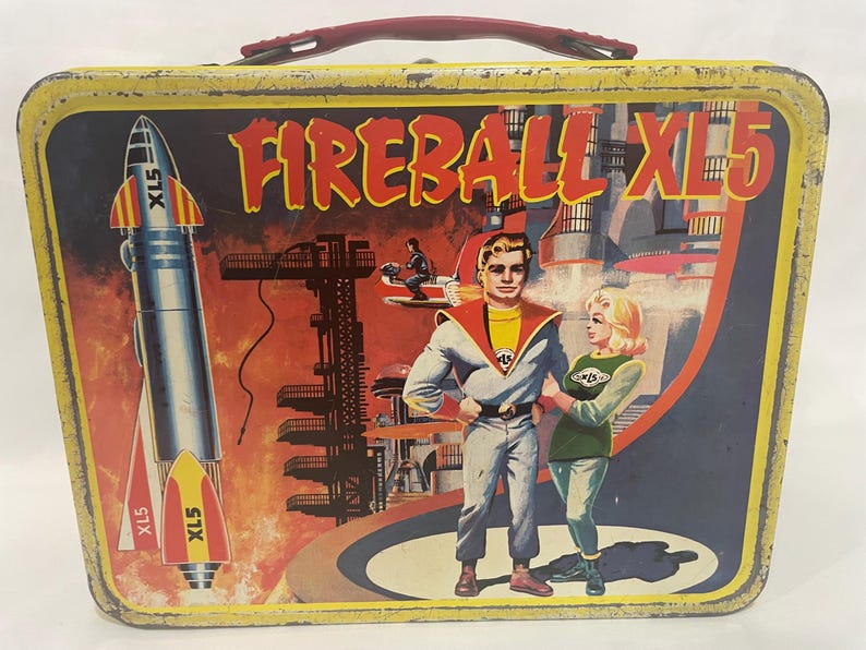 Puede incluir: Fiambrera met&aacute;lica vintage con asa roja y gr&aacute;fico colorido. El gr&aacute;fico presenta el texto "FIREBALL XL5", un cohete y dos personajes en un entorno futurista. La fiambrera tiene un borde amarillo.