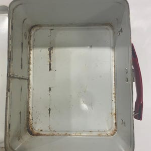 Puede incluir: Una lonchera de metal vintage abierta con un asa roja. El interior es de color gris claro con signos de &oacute;xido y desgaste. La lonchera tiene forma rectangular con esquinas redondeadas.