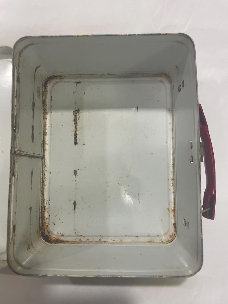 Puede incluir: Una lonchera de metal vintage abierta con un asa roja. El interior es de color gris claro con signos de &oacute;xido y desgaste. La lonchera tiene forma rectangular con esquinas redondeadas.