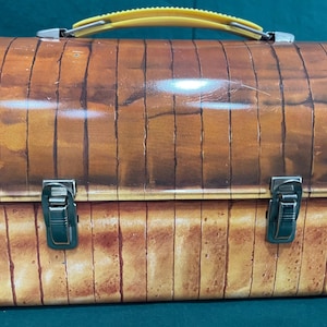 May include: Vintage metal lunchbox with a wood grain design in shades of brown and tan. The lunchbox has a yellow plastic handle and two silver metal clasps. A classic container for carrying meals.