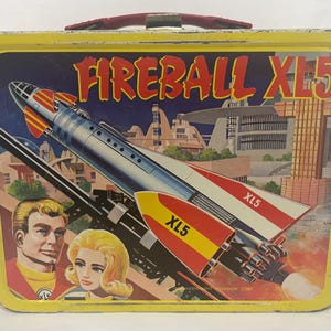 Puede incluir: Fiambrera met&aacute;lica vintage con borde amarillo y asa roja. La parte delantera presenta una colorida ilustraci&oacute;n de un cohete llamado "Fireball XL5" con el texto "XL5" en las alas. La imagen incluye dos personas y un paisaje urbano futurista.