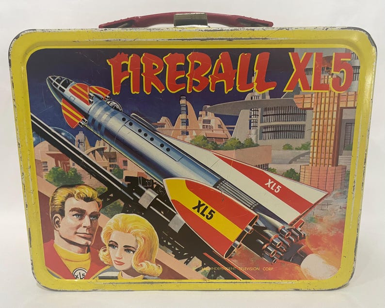 Puede incluir: Fiambrera met&aacute;lica vintage con borde amarillo y asa roja. La parte delantera presenta una colorida ilustraci&oacute;n de un cohete llamado "Fireball XL5" con el texto "XL5" en las alas. La imagen incluye dos personas y un paisaje urbano futurista.