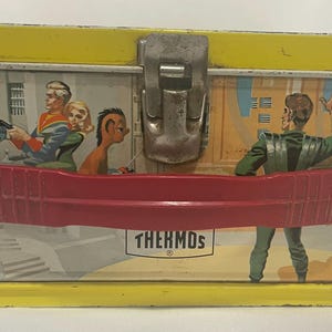 Puede incluir: Lonchera de metal vintage con tapa y base amarillas, con una escena de ciencia ficci&oacute;n retro con figuras con atuendo futurista. La lonchera tiene un asa roja y un cierre de metal. La palabra "THERMOS" est&aacute; impresa en la parte delantera.