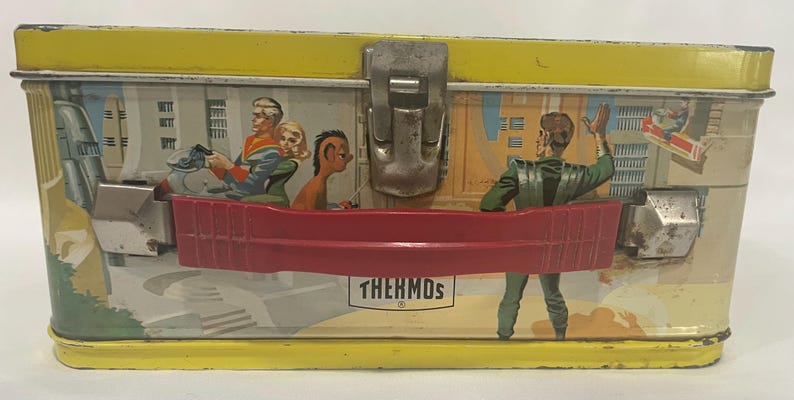 Puede incluir: Lonchera de metal vintage con tapa y base amarillas, con una escena de ciencia ficci&oacute;n retro con figuras con atuendo futurista. La lonchera tiene un asa roja y un cierre de metal. La palabra "THERMOS" est&aacute; impresa en la parte delantera.