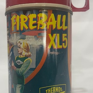 Puede incluir: Termo Fireball XL5 vintage, rojo y verde azulado. El termo tiene una tapa y un asa rojas, con un cuerpo cil&iacute;ndrico. El cuerpo tiene una ilustraci&oacute;n retro de una mujer y el texto "FIREBALL XL5" en amarillo. La palabra "THERMOS" tambi&eacute;n es visible.