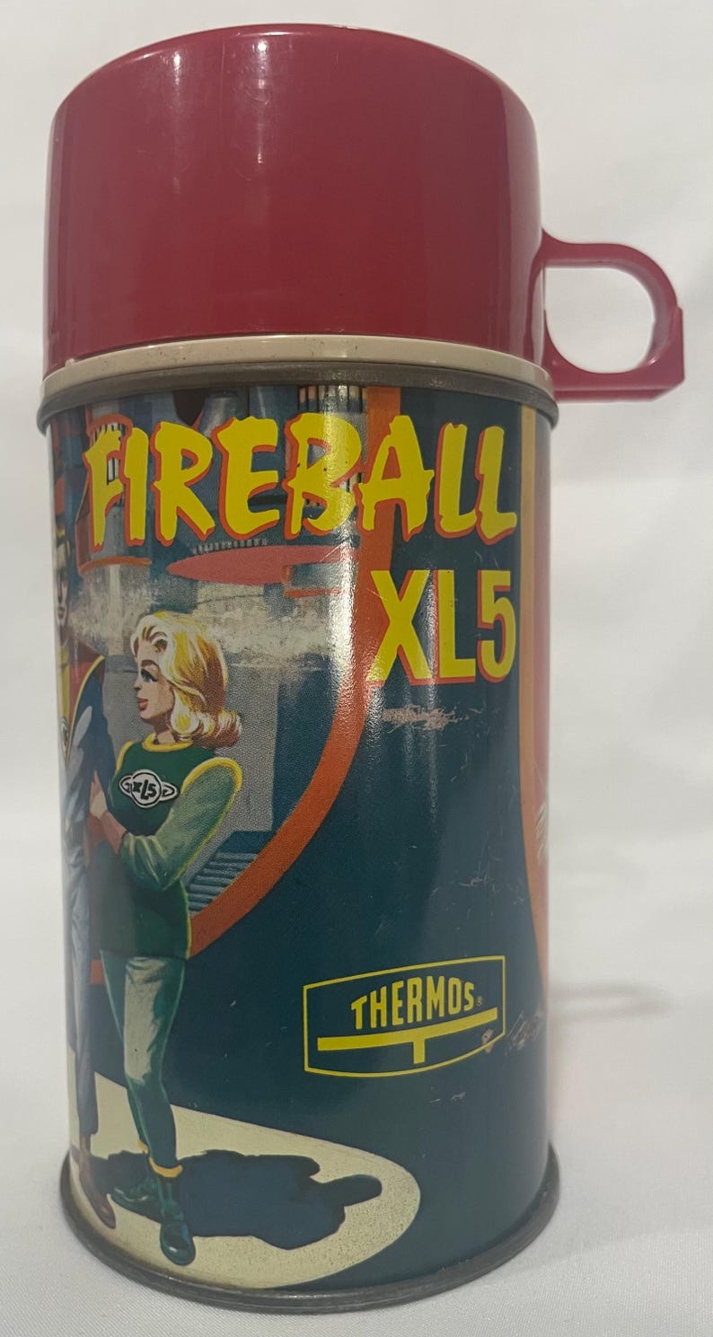 Puede incluir: Termo Fireball XL5 vintage, rojo y verde azulado. El termo tiene una tapa y un asa rojas, con un cuerpo cil&iacute;ndrico. El cuerpo tiene una ilustraci&oacute;n retro de una mujer y el texto "FIREBALL XL5" en amarillo. La palabra "THERMOS" tambi&eacute;n es visible.