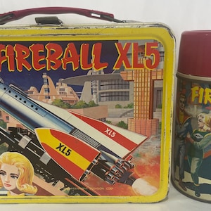 Puede incluir: Juego vintage de fiambrera y termo Fireball XL5. La fiambrera es amarilla con asa roja y presenta un gr&aacute;fico de cohete. El termo es granate y verde azulado con un gr&aacute;fico a juego. Un coleccionable retro.