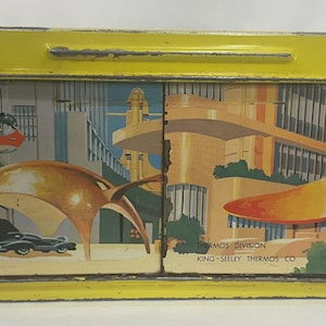 Puede incluir: Fiambrera vintage de metal amarillo con asa. La fiambrera presenta una colorida ilustraci&oacute;n de una ciudad futurista con veh&iacute;culos voladores y arquitectura moderna. El texto "THERMOS DIVISION KING-SEELEY THERMOS CO" est&aacute; impreso en el lateral.