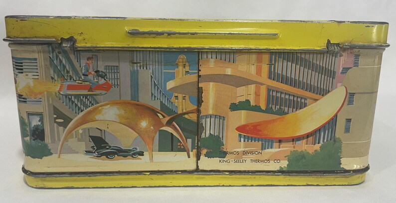 Puede incluir: Fiambrera vintage de metal amarillo con asa. La fiambrera presenta una colorida ilustraci&oacute;n de una ciudad futurista con veh&iacute;culos voladores y arquitectura moderna. El texto "THERMOS DIVISION KING-SEELEY THERMOS CO" est&aacute; impreso en el lateral.