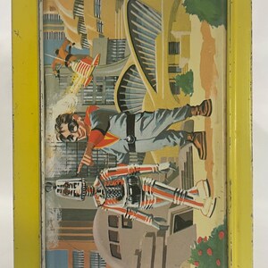 Puede incluir: Fiambrera met&aacute;lica vintage con borde amarillo. La ilustraci&oacute;n representa un paisaje urbano futurista con un hombre con mono gris y un robot. Una persona con un jetpack vuela en el fondo. La fiambrera tiene un dise&ntilde;o retro de mediados de siglo.