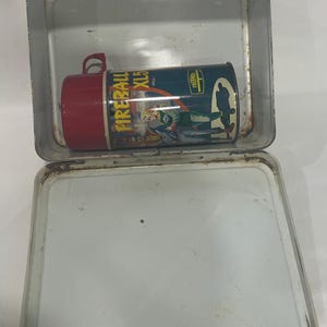 Puede incluir: Lonchera met&aacute;lica vintage con asa roja y termo a juego. El termo tiene tapa roja y presenta el texto "FIREBALL XL5" y una imagen de dibujos animados. La lonchera est&aacute; abierta, mostrando el interior.