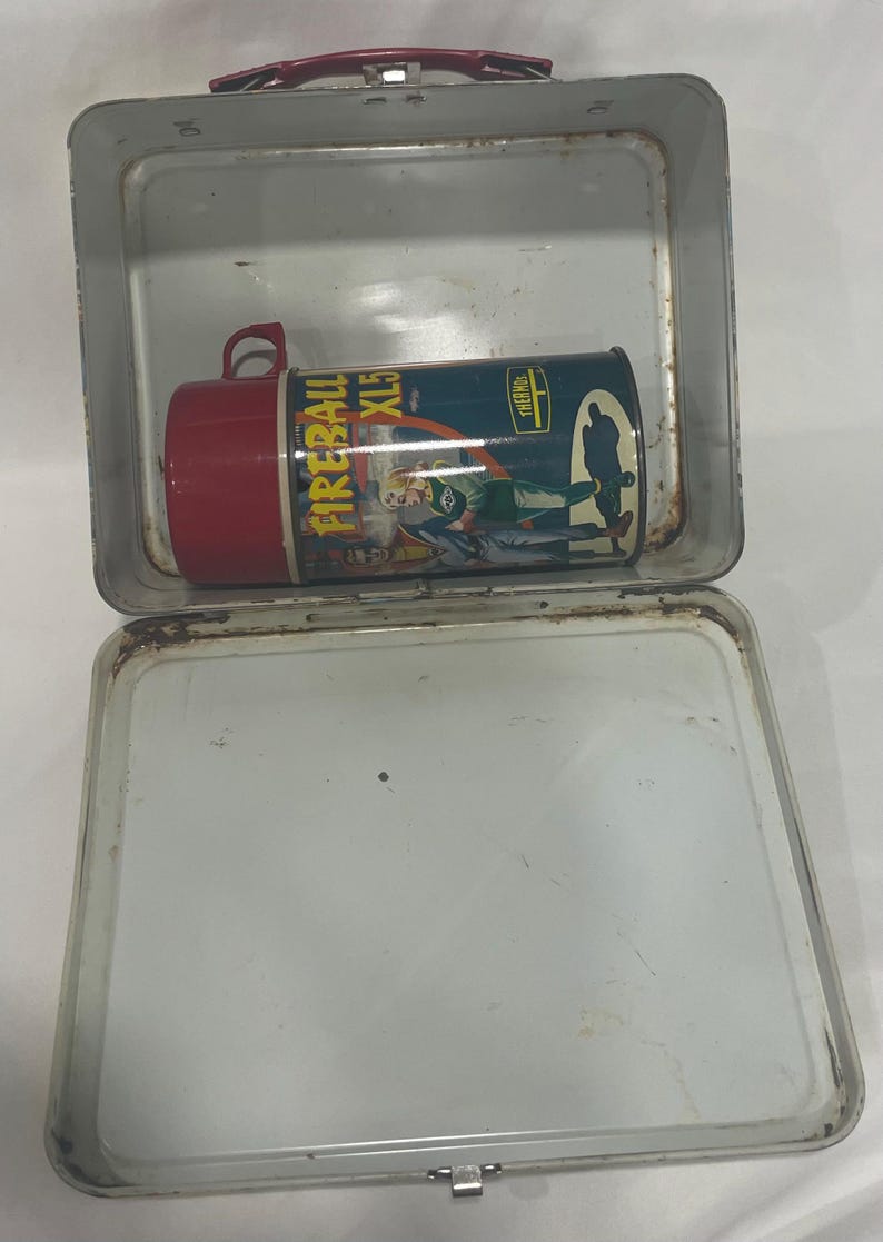 Puede incluir: Lonchera met&aacute;lica vintage con asa roja y termo a juego. El termo tiene tapa roja y presenta el texto "FIREBALL XL5" y una imagen de dibujos animados. La lonchera est&aacute; abierta, mostrando el interior.