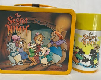 May include: Yellow lunchbox and matching thermos featuring characters from "The Secret of NIMH." The lunchbox has a hinged lid and a metal clasp. The thermos has a yellow lid and handle. Both items display colorful illustrations.