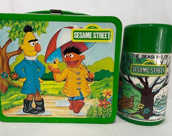 Può includere: Set vintage verde di lunch box e thermos di Sesame Street. Il lunch box presenta Ernie e Bert sotto la pioggia. Il thermos ha coperchio e manico verdi, con una scena invernale e la scritta "The Seasons of Sesame Street."