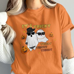 May include: Orange t-shirt with a Halloween-themed graphic. The design features two cartoon ghosts wearing sunglasses and a gold chain, with the text "IT'S TRICKY" and "TRICKY TRICKY TRICKY". Pumpkins and stars complete the design.