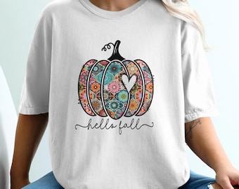 May include: White t-shirt featuring a colorful pumpkin design with floral patterns and a white heart. The words "hello fall" are written in a script font below the pumpkin. The shirt is a casual, fall-themed apparel item.