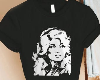 May include: A black short-sleeved t-shirt featuring a high-contrast white and grey graphic print of a person with voluminous hair. The design is centered on the chest area of the garment, which is displayed on a wooden hanger against a neutral background.