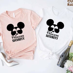 May include: Two short-sleeved t-shirts, one light pink and one white, each featuring a black graphic of a mouse head wearing sunglasses. The text "PRINCESS SECURITY" is printed below the graphic. The shirts are part of a gift set.