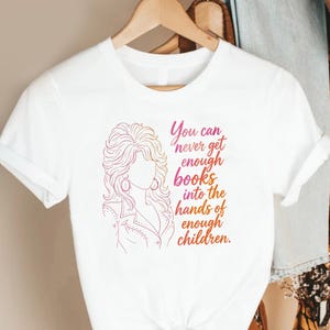 May include: A white short-sleeved t-shirt featuring a pink and orange gradient line art illustration of a person with voluminous hair and the text: You can never get enough books into the hands of enough children. The shirt is displayed on a hanger.