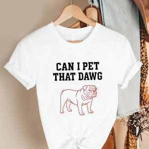 May include: White t-shirt with the text "CAN I PET THAT DAWG" in black, above a red outline of a bulldog. The shirt is hanging on a wooden hanger.