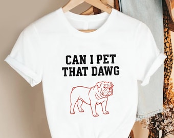 May include: White t-shirt with the text "CAN I PET THAT DAWG" in black, above a red outline of a bulldog. The shirt is hanging on a wooden hanger.