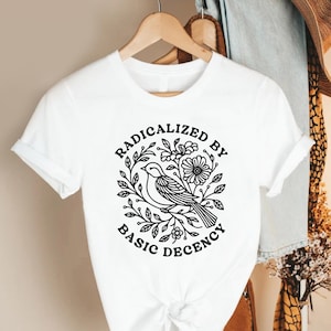 May include: White t-shirt featuring a black and white graphic of a bird surrounded by leaves and flowers. The text "RADICALIZED BY BASIC DECENCY" is printed around the bird design. The shirt is hanging on a wooden hanger.
