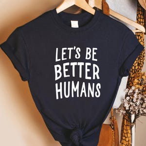 May include: A navy blue t-shirt with the white text "LET'S BE BETTER HUMANS." The shirt is hanging on a wooden hanger. The sleeves are cuffed. The shirt is knotted at the bottom.