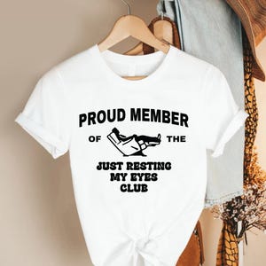 May include: White t-shirt with the text "PROUD MEMBER OF THE JUST RESTING MY EYES CLUB" in black. The graphic shows a person reclining in a chair. The shirt is hanging on a wooden hanger.