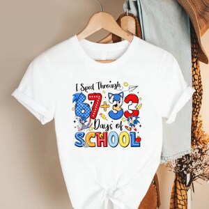 May include: White t-shirt featuring a colorful graphic with the text "I sped through 67 + 33 Days of SCHOOL". The design includes cartoon characters and numbers in blue, red, and yellow.