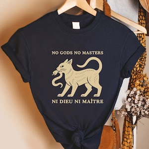 May include: Navy blue t-shirt with the text "NO GODS NO MASTERS" above a tan cat holding a snake in its mouth. Below the cat is the text "NI DIEU NI MAÎTRE". The shirt is hanging on a wooden hanger.