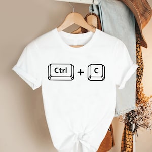 May include: White t-shirt featuring a black graphic of the Ctrl + C keyboard shortcut. The design includes the word "Ctrl" inside a rectangle, followed by a plus sign, and the letter "C" inside a rectangle. The shirt is short-sleeved.