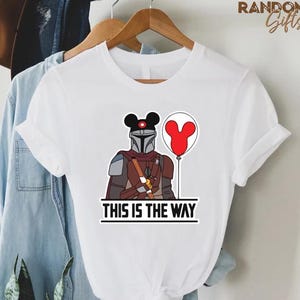 Puede incluir: Camiseta blanca con una ilustraci&oacute;n de dibujos animados de un personaje mandaloriano con una cabeza de Mickey Mouse y un globo rojo. El texto "THIS IS THE WAY" est&aacute; impreso debajo. La camiseta est&aacute; colgada en una percha de madera.