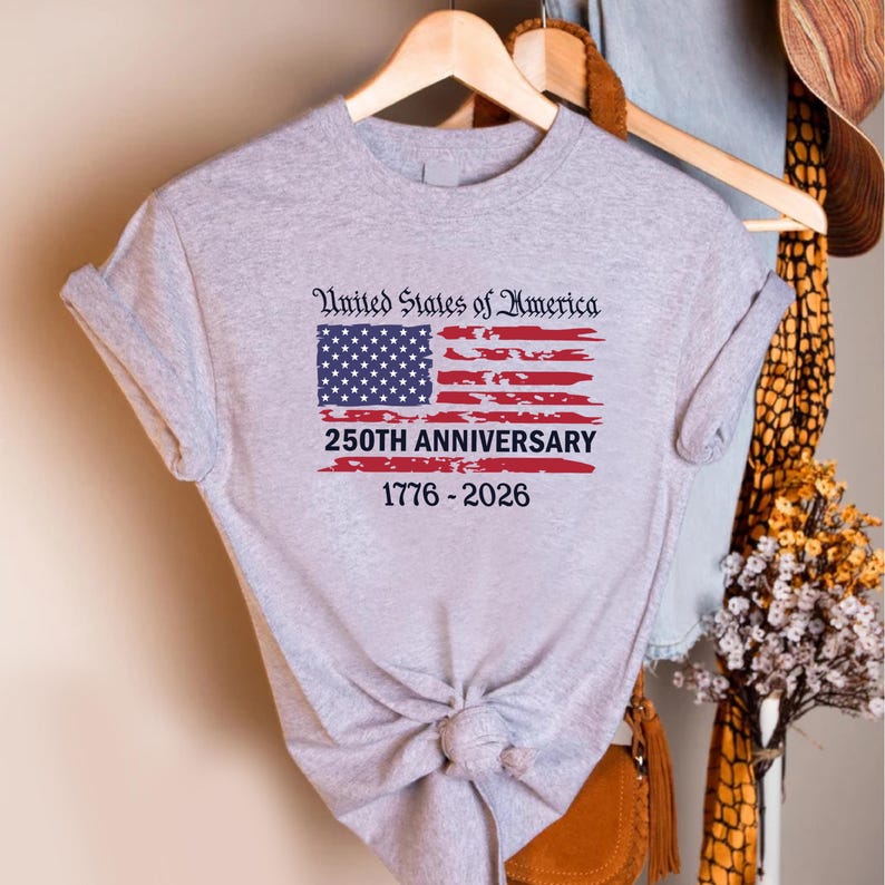 May include: A light gray t-shirt featuring a distressed American flag design. The flag includes the text "United States of America" above the flag, and "250th Anniversary 1776-2026" below. The shirt is hanging on a wooden hanger.