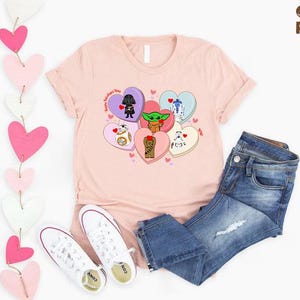 May include: A light pink t-shirt featuring cartoon characters on heart-shaped candies. The shirt is paired with blue jeans and white sneakers. A string of pink and white hearts hangs on the left side. The text "Custom Random Gifts" is in the upper right corner.