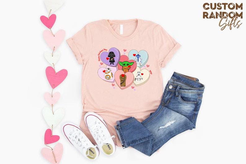 May include: A light pink t-shirt featuring cartoon characters on heart-shaped candies. The shirt is paired with blue jeans and white sneakers. A string of pink and white hearts hangs on the left side. The text "Custom Random Gifts" is in the upper right corner.