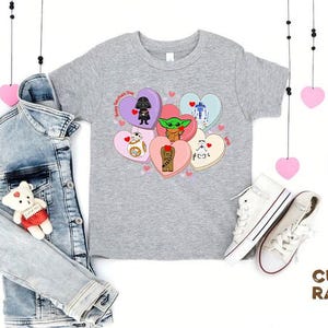 May include: A gray t-shirt featuring Star Wars characters on Valentine's Day heart-shaped candies. A denim jacket, white sneakers, and a teddy bear are also in the image. Pink heart decorations hang from above.
