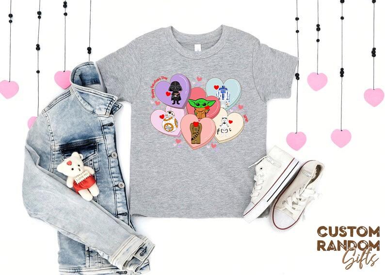 May include: A gray t-shirt featuring Star Wars characters on Valentine's Day heart-shaped candies. A denim jacket, white sneakers, and a teddy bear are also in the image. Pink heart decorations hang from above.
