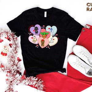 May include: Black t-shirt featuring Star Wars characters on heart-shaped candies. The design includes Darth Vader, Baby Yoda, R2-D2, BB-8, Chewbacca, and a Stormtrooper. Paired with red pants and white sneakers, with a Valentine's Day wreath.